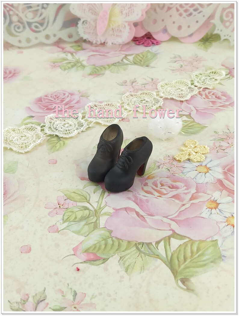 May include: A pair of miniature black lace-up high-heeled shoes with rounded toes and red soles. The shoes feature a smooth matte finish and are positioned on a surface with a floral pattern. The text "The hand flower" is visible in the center.