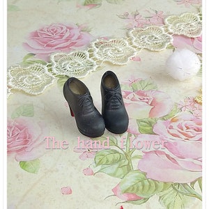 May include: A pair of miniature black lace-up high-heeled shoes with red soles. The shoes feature a rounded toe, a structured heel, and visible laces on the front. The text "The hand flower" is printed in light pink letters across the center.