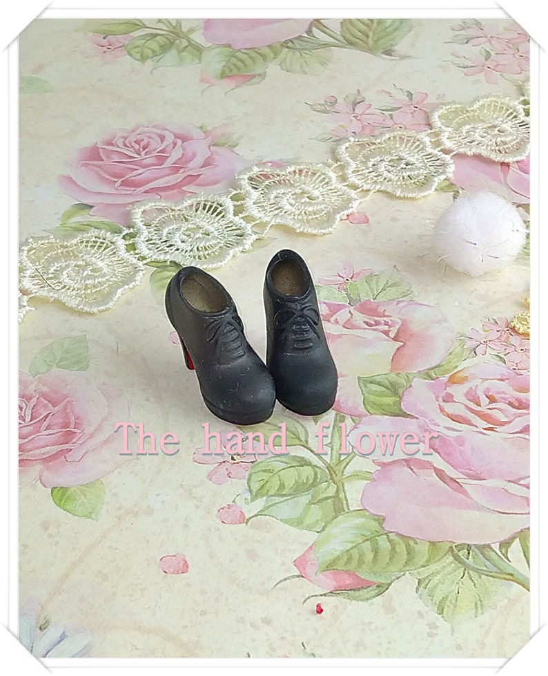 May include: A pair of miniature black lace-up high-heeled shoes with red soles. The shoes feature a rounded toe, a structured heel, and visible laces on the front. The text "The hand flower" is printed in light pink letters across the center.