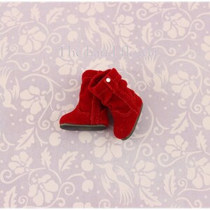 Puede incluir: Un par de botas de ante rojo con un dobladillo y un peque&ntilde;o bot&oacute;n blanco. Las botas est&aacute;n sobre un fondo blanco con un patr&oacute;n floral.