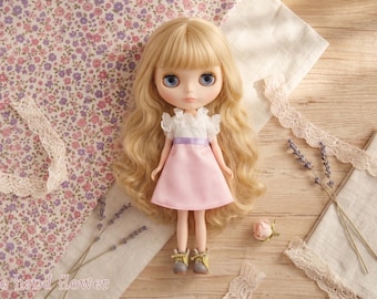 May include: A Blythe doll with long, wavy blonde hair and blue eyes, wearing a white top with a pink dress and gray boots. The doll is surrounded by floral fabric, lace, lavender sprigs, and a rose on a wooden surface.