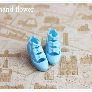 Op de afbeelding: Een paar lichtblauwe poppen schoenen met strikken. De schoenen liggen op een achtergrond van een bedrukte kaart.