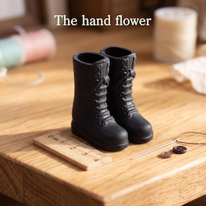 May include: A pair of miniature black lace-up boots positioned next to a small wooden ruler showing a length of approximately 3 centimeters. The text The hand flower is visible above the boots, accompanied by a needle, thread, and buttons.