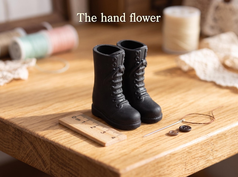 May include: A pair of miniature black lace-up boots positioned next to a small wooden ruler showing a length of approximately 3 centimeters. The text The hand flower is visible above the boots, accompanied by a needle, thread, and buttons.