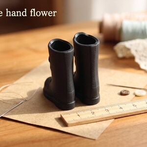 May include: A pair of miniature black boots with a smooth finish, positioned next to a wooden ruler showing measurements in centimeters. The text 'The hand flower' is visible in the upper left corner, accompanied by sewing needles and buttons.