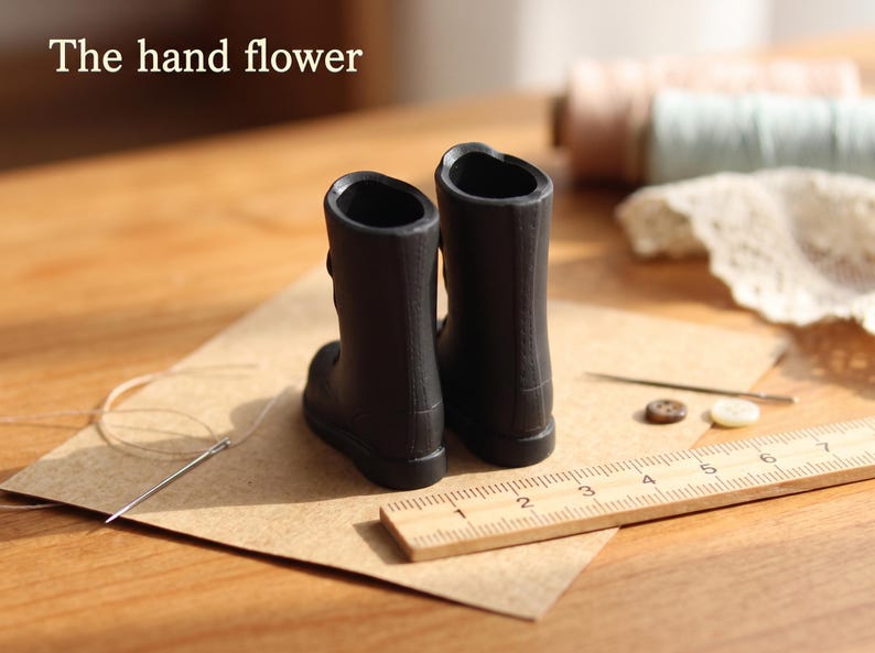May include: A pair of miniature black boots with a smooth finish, positioned next to a wooden ruler showing measurements in centimeters. The text 'The hand flower' is visible in the upper left corner, accompanied by sewing needles and buttons.