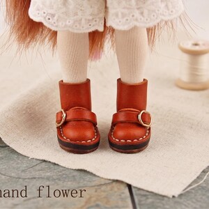 May include: A pair of miniature brown leather boots with gold-colored buckles. The boots are stitched with white thread and have a dark brown sole. The text "The hand flower" is visible at the bottom of the image.