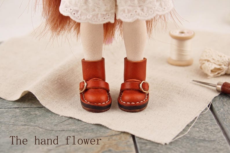 May include: A pair of miniature brown leather boots with gold-colored buckles. The boots are stitched with white thread and have a dark brown sole. The text "The hand flower" is visible at the bottom of the image.