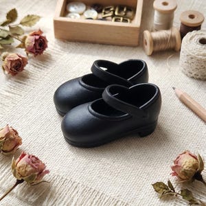 Pode incluir: Um par de sapatos Mary Jane pretos com biqueiras arredondadas e uma tira sobre o peito do pé. Os sapatos estão posicionados perto de uma caixa de madeira com botões, vários carretéis de linha de madeira, um lápis e rosas secas.