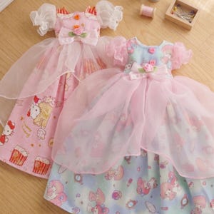May include: Two doll dresses with pink and white accents. One dress features a pink bodice with a bow and a floral embellishment, and a skirt with a Hello Kitty print. The other dress has a blue bodice and a skirt with a cartoon character print.