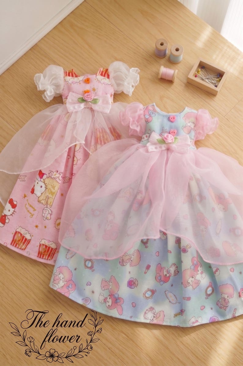 May include: Two doll dresses with pink and white accents. One dress features a pink bodice with a bow and a floral embellishment, and a skirt with a Hello Kitty print. The other dress has a blue bodice and a skirt with a cartoon character print.