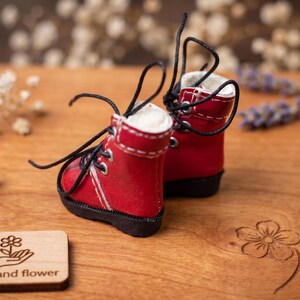 May include: A pair of miniature red boots with black laces and white stitching. The boots are set on a wooden surface with a small wooden tag that reads "The hand flower". Decorative flowers and foliage surround the boots.