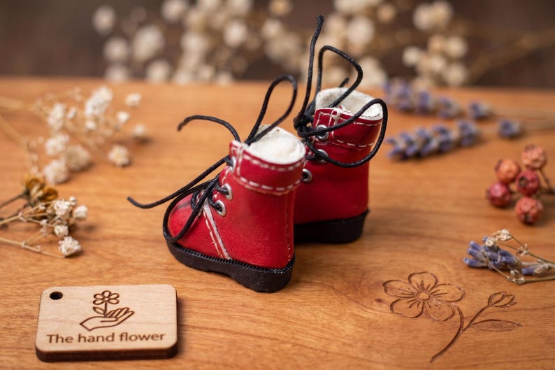 May include: A pair of miniature red boots with black laces and white stitching. The boots are set on a wooden surface with a small wooden tag that reads "The hand flower". Decorative flowers and foliage surround the boots.