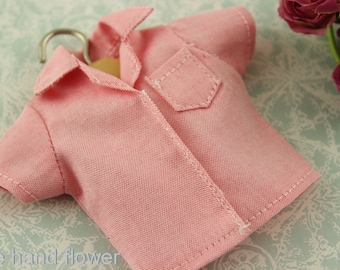 May include: A miniature pink button-down shirt with a small pocket and white stitching details. The shirt is hanging on a wooden hanger with a silver hook. The fabric has a subtle texture. The words "The hand flower" are visible.