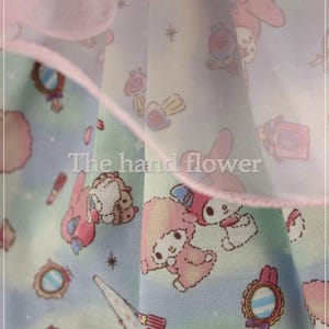 May include: A light blue fabric with a repeating pattern of cartoon characters, mirrors, and makeup items. The fabric has a pink ruffle trim and the text "The hand flower".