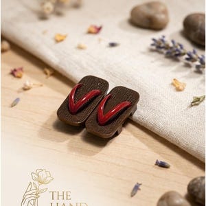 May include: A pair of miniature wooden geta sandals with red straps. The sandals are brown with a dark red thong. The sandals are displayed on a light wooden surface with decorative elements, including dried flowers and stones. The logo "THE HAND FLOWER" is visible.