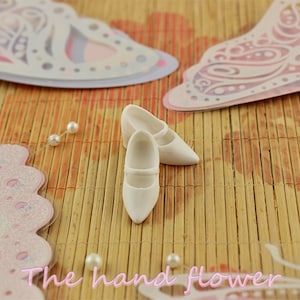 May include: Two white doll shoes with a strap across the top, sitting on a woven bamboo mat with pink and white butterfly cutouts.