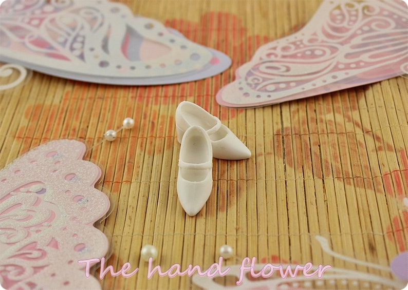 May include: Two white doll shoes with a strap across the top, sitting on a woven bamboo mat with pink and white butterfly cutouts.