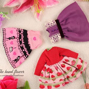 May include: A collection of doll clothes, including a pink dress with black cat and heart patterns, a purple dress with lace trim, and a red dress with strawberry designs. The hand flower Etsy shop logo is visible.