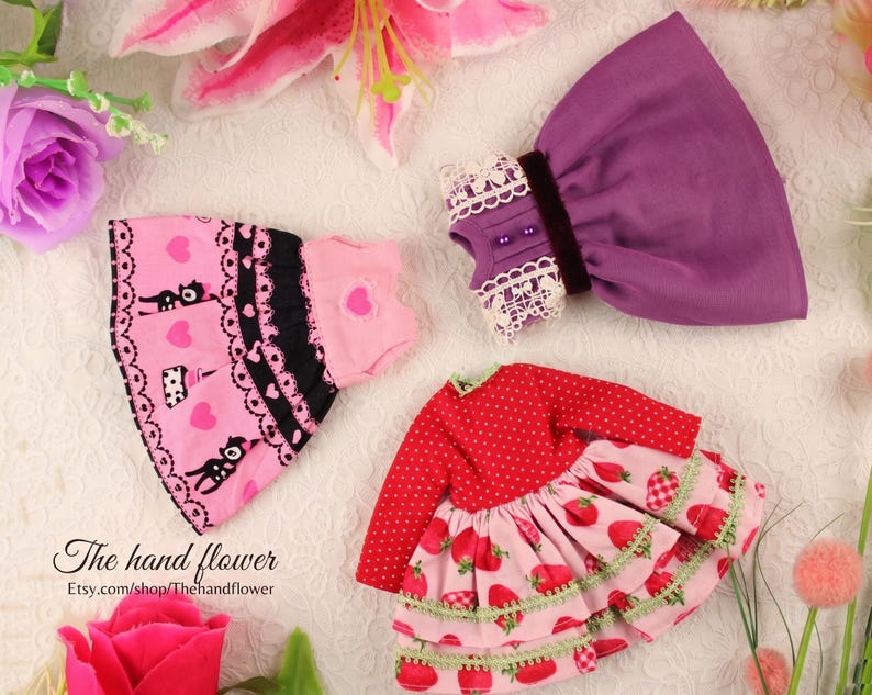 May include: A collection of doll clothes, including a pink dress with black cat and heart patterns, a purple dress with lace trim, and a red dress with strawberry designs. The hand flower Etsy shop logo is visible.