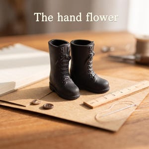 May include: A pair of miniature black lace-up boots with a matte finish, positioned next to a small wooden ruler showing centimeter markings, two brown buttons, and a needle with thread. The text "The hand flower" is visible above.