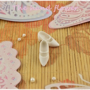 May include: Two white doll shoes with a closed toe and a small heel. The shoes are on a light brown bamboo mat with pink and white butterfly decorations.