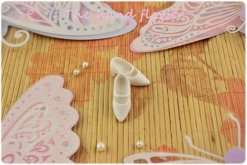 May include: Two white doll shoes with a closed toe and a small heel. The shoes are on a light brown bamboo mat with pink and white butterfly decorations.
