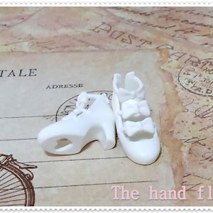 Op de afbeelding: Een paar witte miniatuur pumps met strikaccenten. De schoenen staan op een vintage ansichtkaart achtergrond, met de tekst "The hand flower" onderaan.