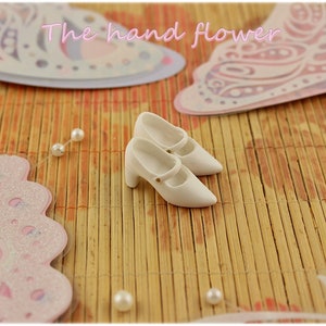 May include: A pair of white doll shoes with a strap and a small heel. The shoes are on a bamboo mat with pink butterfly decorations.