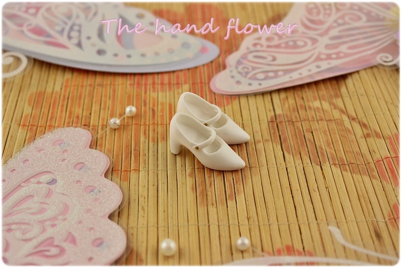 May include: A pair of white doll shoes with a strap and a small heel. The shoes are on a bamboo mat with pink butterfly decorations.