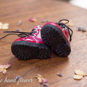 May include: A pair of small, red boots with black laces and soles. The boots are set on a wooden surface, surrounded by dried flowers and petals. The boots have a glossy finish.