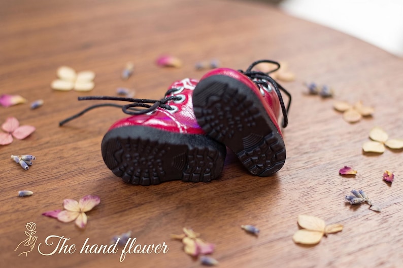 May include: A pair of small, red boots with black laces and soles. The boots are set on a wooden surface, surrounded by dried flowers and petals. The boots have a glossy finish.