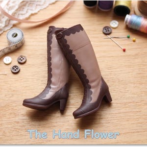 May include: A pair of miniature knee-high boots featuring a two-tone design with light tan shafts and dark brown lower sections, heels, and scalloped trim along the front. The text The Hand Flower is printed at the bottom of the image.