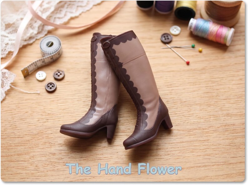 May include: A pair of miniature knee-high boots featuring a two-tone design with light tan shafts and dark brown lower sections, heels, and scalloped trim along the front. The text The Hand Flower is printed at the bottom of the image.