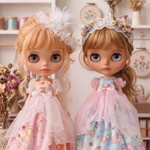 May include: Two dolls with blonde hair and large eyes are dressed in pink and blue dresses. The doll on the left wears a white headpiece, and the doll on the right wears a floral crown. The dresses have cartoon character prints. The text "The Hand Flower" is at the bottom.