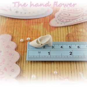 May include: A white ceramic shoe with a gold buckle, measuring approximately 1 inch in length, is placed on a blue ruler with black markings.