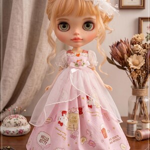 May include: A doll with blonde hair and green eyes wearing a pink dress with a Hello Kitty print. The doll has a white feathered headpiece. The dress has a sheer overlay. The text "The hand flower" is at the bottom of the image.