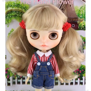 May include: A doll with blonde hair styled in pigtails, held by red scrunchies. The doll wears a red and white striped shirt with a white collar, denim overalls, and has large brown eyes. The text "The Hand Flower" is at the top.