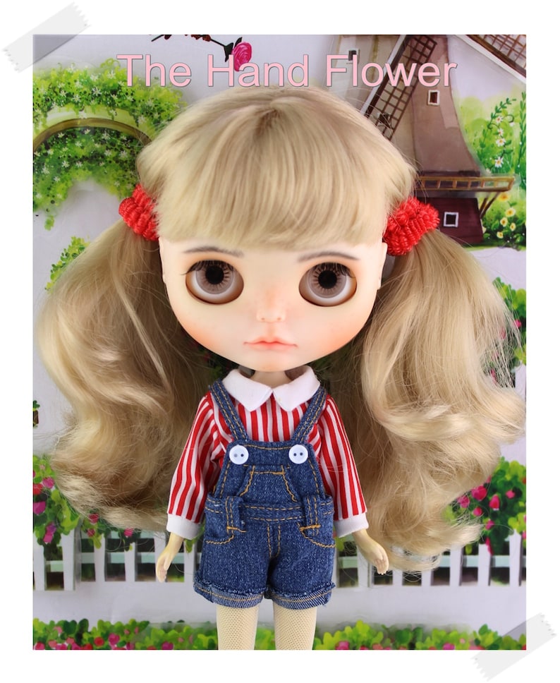 May include: A doll with blonde hair styled in pigtails, held by red scrunchies. The doll wears a red and white striped shirt with a white collar, denim overalls, and has large brown eyes. The text "The Hand Flower" is at the top.