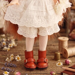 May include: A doll wearing a white dress with lace trim, white shorts, and brown leather boots with gold buckles. The doll is standing on a wooden surface with flowers and sewing supplies. The text "The hand flower" is at the bottom.