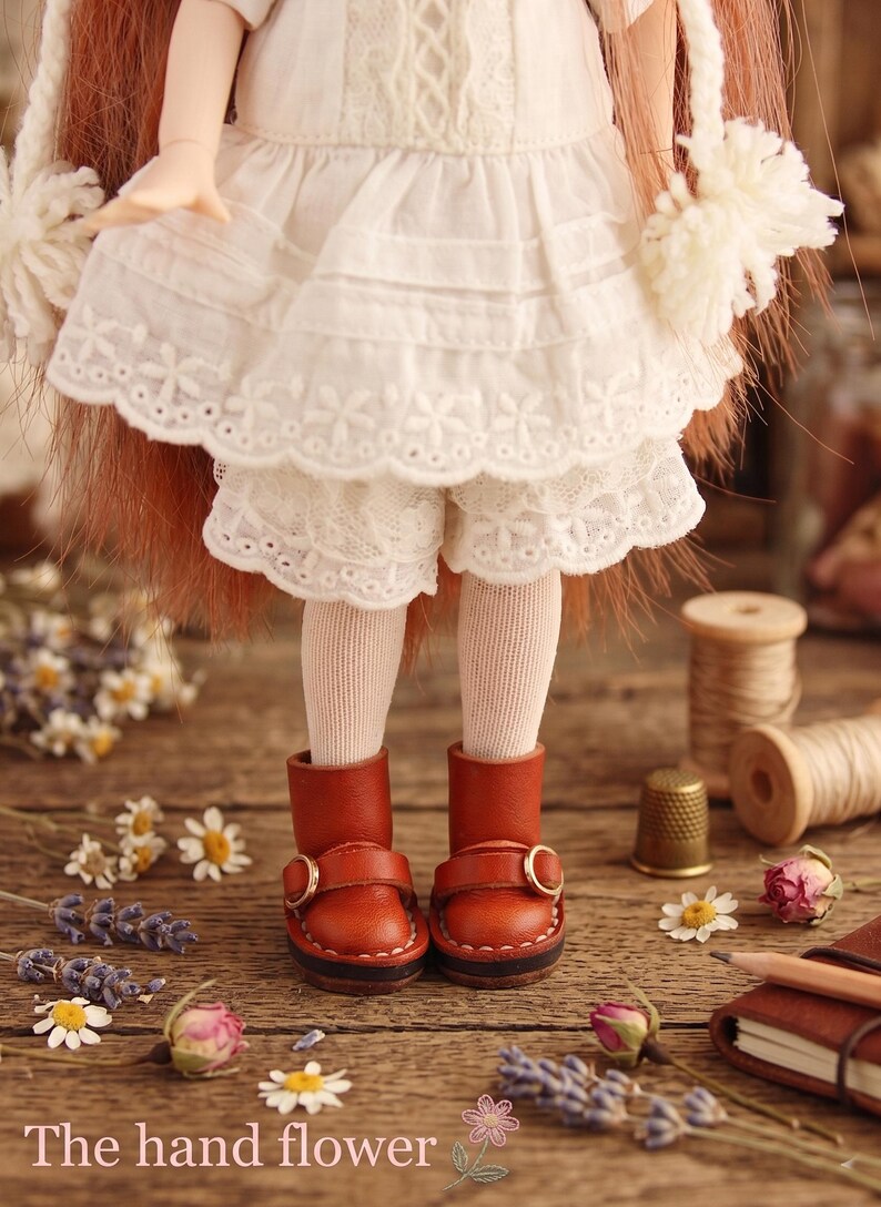 May include: A doll wearing a white dress with lace trim, white shorts, and brown leather boots with gold buckles. The doll is standing on a wooden surface with flowers and sewing supplies. The text "The hand flower" is at the bottom.