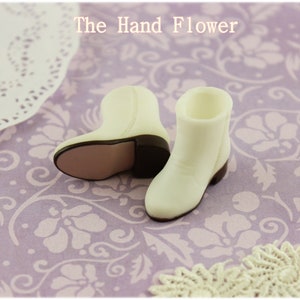 Puede incluir: Un par de botas de vaquero blancas en miniatura con tacones y suelas marrones. Las botas est&aacute;n sobre una superficie con estampado floral lavanda. El texto "The Hand Flower" es visible en la parte superior.