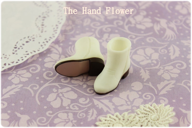 Puede incluir: Un par de botas de vaquero blancas en miniatura con tacones y suelas marrones. Las botas est&aacute;n sobre una superficie con estampado floral lavanda. El texto "The Hand Flower" es visible en la parte superior.