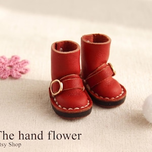 May include: A pair of miniature red leather boots with gold buckles, featuring white stitching around the soles. A pink flower and a white pom-pom are also in the image. The Etsy shop name "The hand flower" is visible.