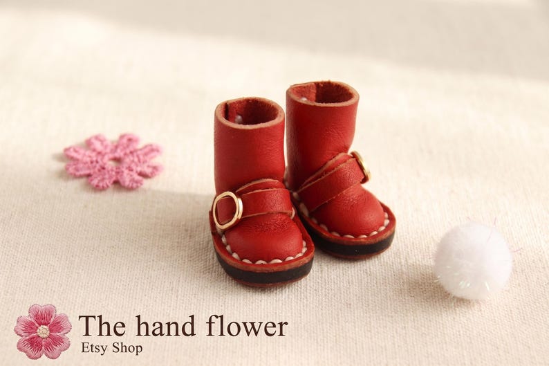 May include: A pair of miniature red leather boots with gold buckles, featuring white stitching around the soles. A pink flower and a white pom-pom are also in the image. The Etsy shop name "The hand flower" is visible.