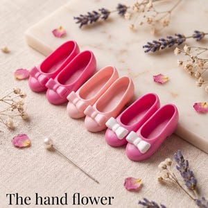 May include: Three pairs of miniature doll shoes in shades of hot pink and light pink, each featuring a small bow on the toe. A single pearl-topped sewing pin lies nearby. The text "The hand flower" is printed in black at the bottom of the image.