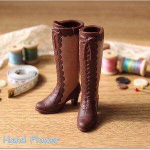 May include: A pair of miniature brown boots with a two-tone design, featuring dark brown lace-up fronts, scalloped edges, and small heels. The boots are crafted with a smooth finish and include decorative bow details at the top of the shafts.