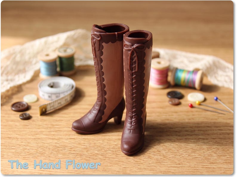 May include: A pair of miniature brown boots with a two-tone design, featuring dark brown lace-up fronts, scalloped edges, and small heels. The boots are crafted with a smooth finish and include decorative bow details at the top of the shafts.