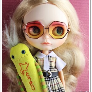 May include: A doll with blonde hair wearing a plaid dress and sunglasses. The doll is holding a yellow plush toy with green and brown stripes. The toy has the word "Grandpa" embroidered on it.