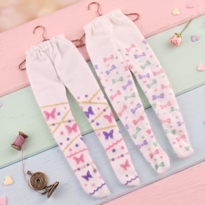 May include: Two pairs of white doll leggings with colorful butterfly and bow patterns, hanging on copper hangers. One pair features pink and purple butterflies, the other has bows in pink, green, and purple. The text "The Hand Flower" is visible.